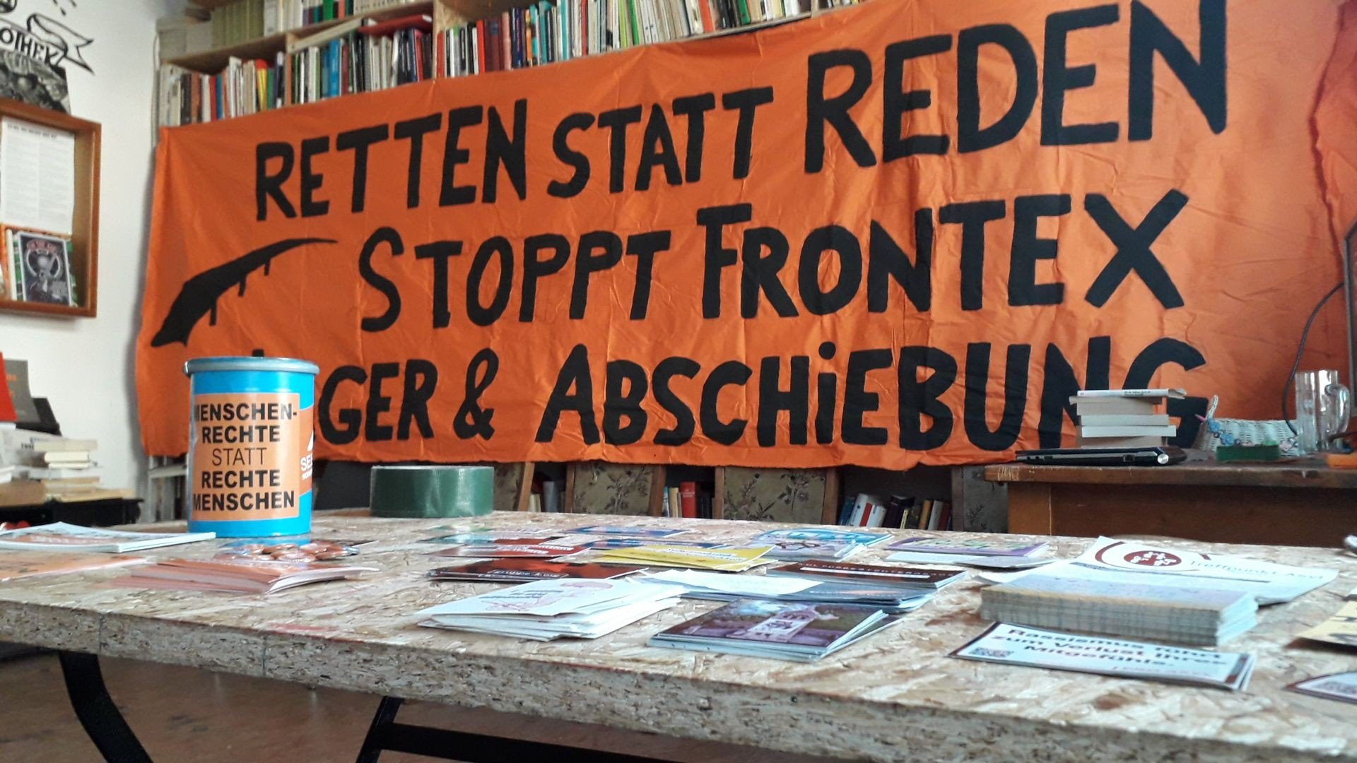 orangenes Transparent mit Aufschrift „Retten statt Reden – Stoppt Frontex, Lager & Abschiebung“, davor ein Tisch mit Infomaterialien und einer Spendenbox; hinter dem Transparent ist ein Bücherregal zu sehen (Gustav-Landauer-Bibliothek Witten)