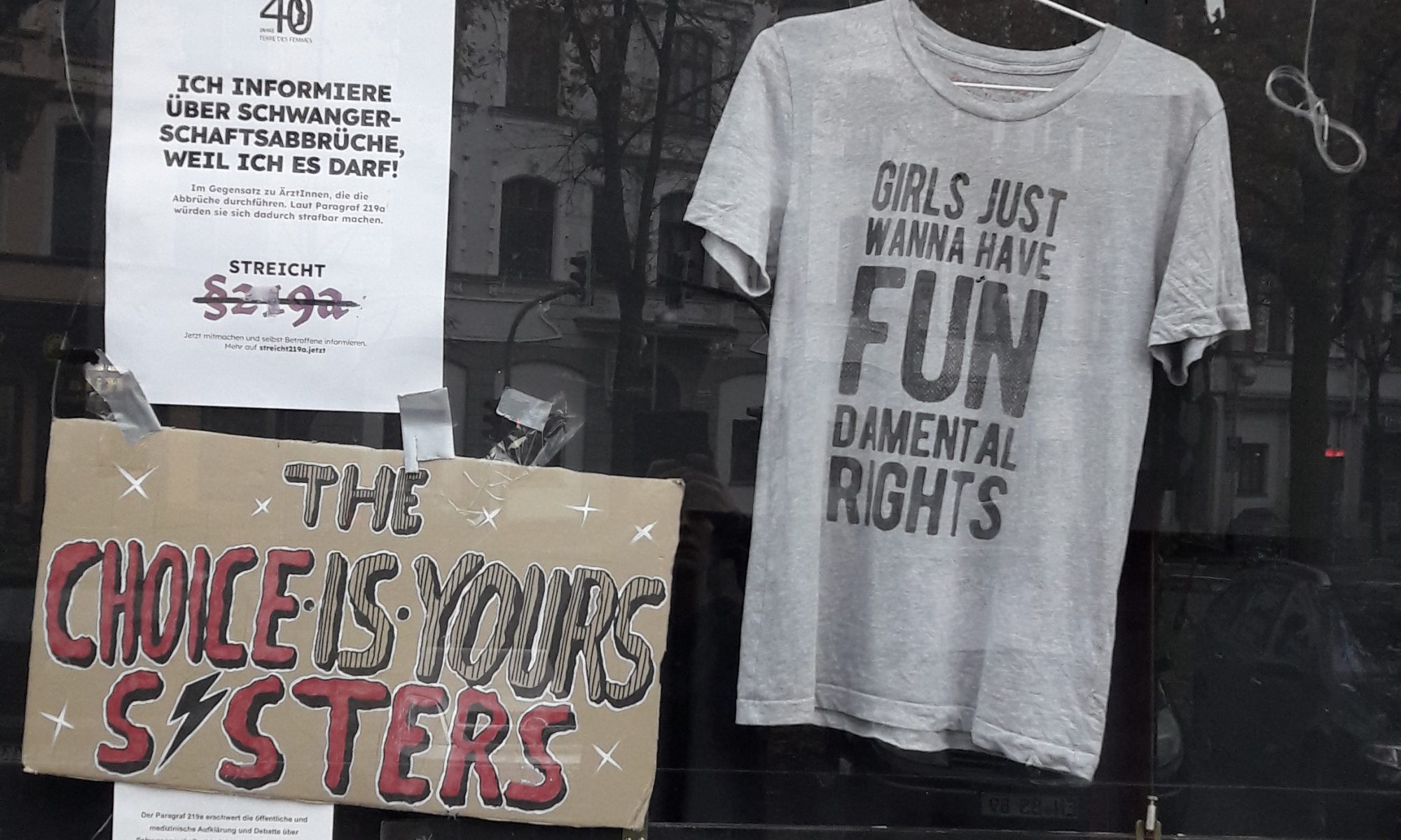 Schaufenster zum Thema Paragraf 219a: ein T-Shirt, eine Tafel „The choice is yours, sisters“, fünf A4-Zettel und ein A3-Zettel mit Texten