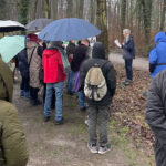 Eine Gruppe Menschen mit Regenschirmen steht am Rand eines Waldes und hört einem älteren Mann zu, der von einem Blatt Papier etwas vorträgt.
