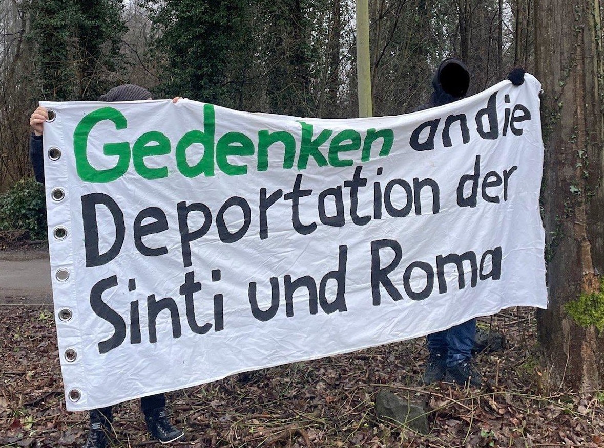 Zwei Personen im Wald halten ein weißes Transparent mit Aufschrift „Gedenken an die Deportation der Sinti und Roma“.