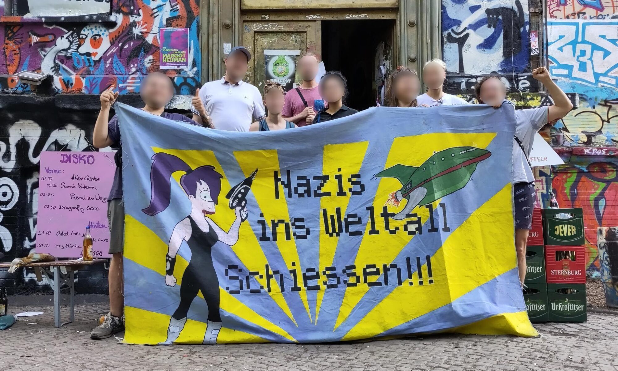 Eine Gruppe Personen mit unkenntlich gemachten Gesichtern hinter einem Transparent mit Aufschrift „Nazis ins Weltall schießen”. Hinter ihnen ein Haus mit vielen Graffiti (Reilstraße 78 in Halle (Saale)).